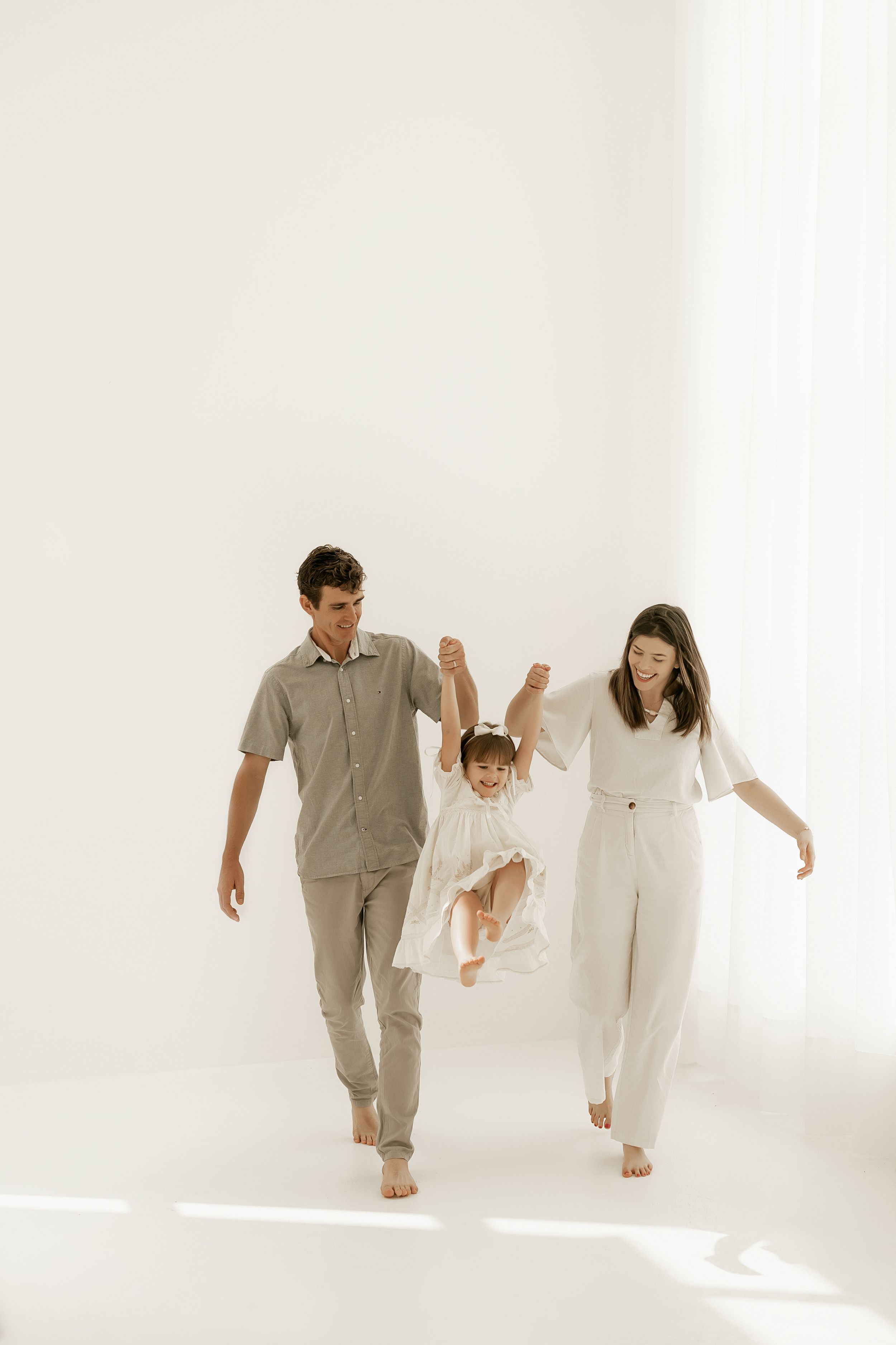Family in casual neutrals swings child while walking together in bright white studio.