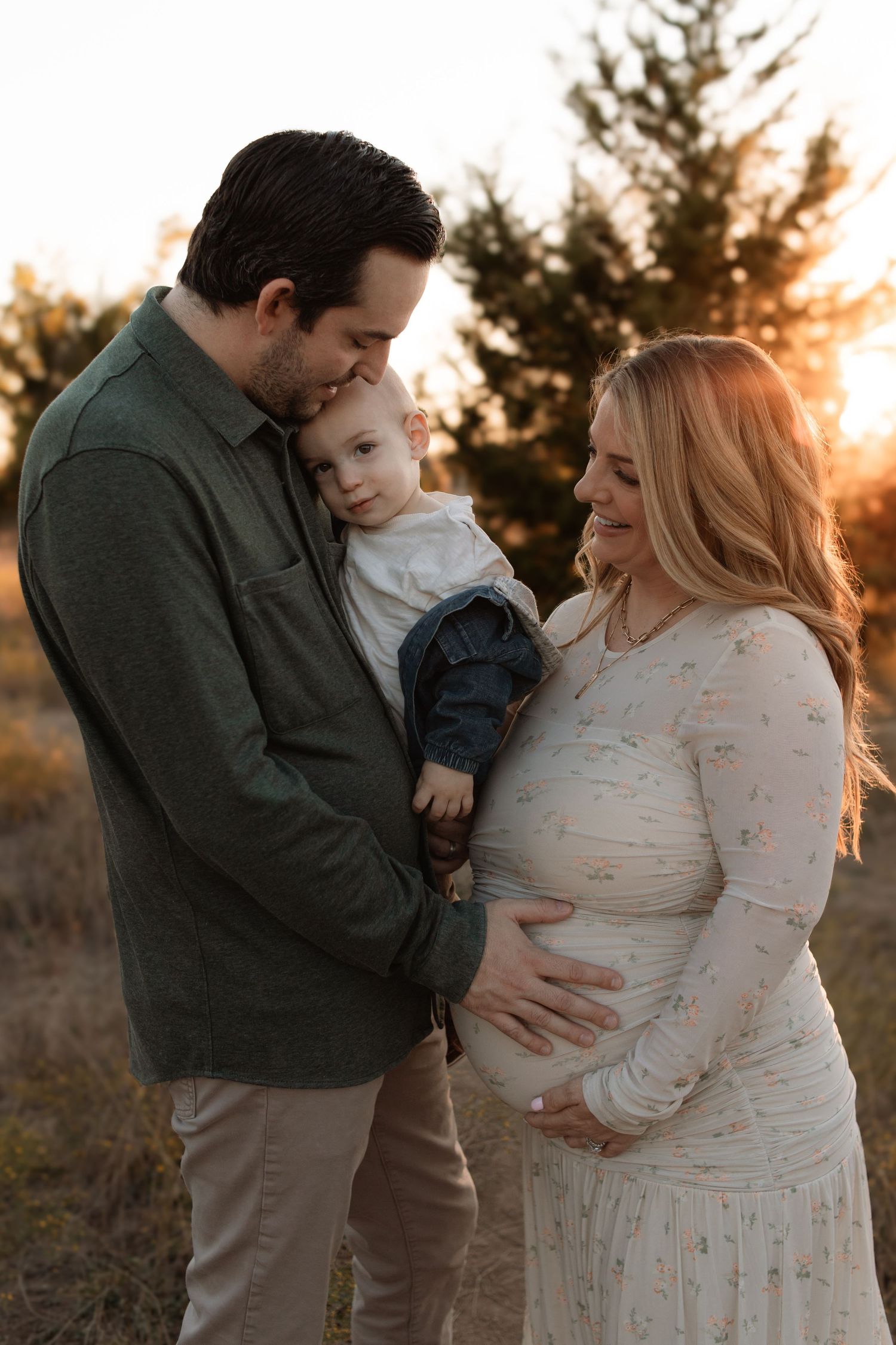 Golden sunset maternity photo with family embracing outdoors among pine trees.