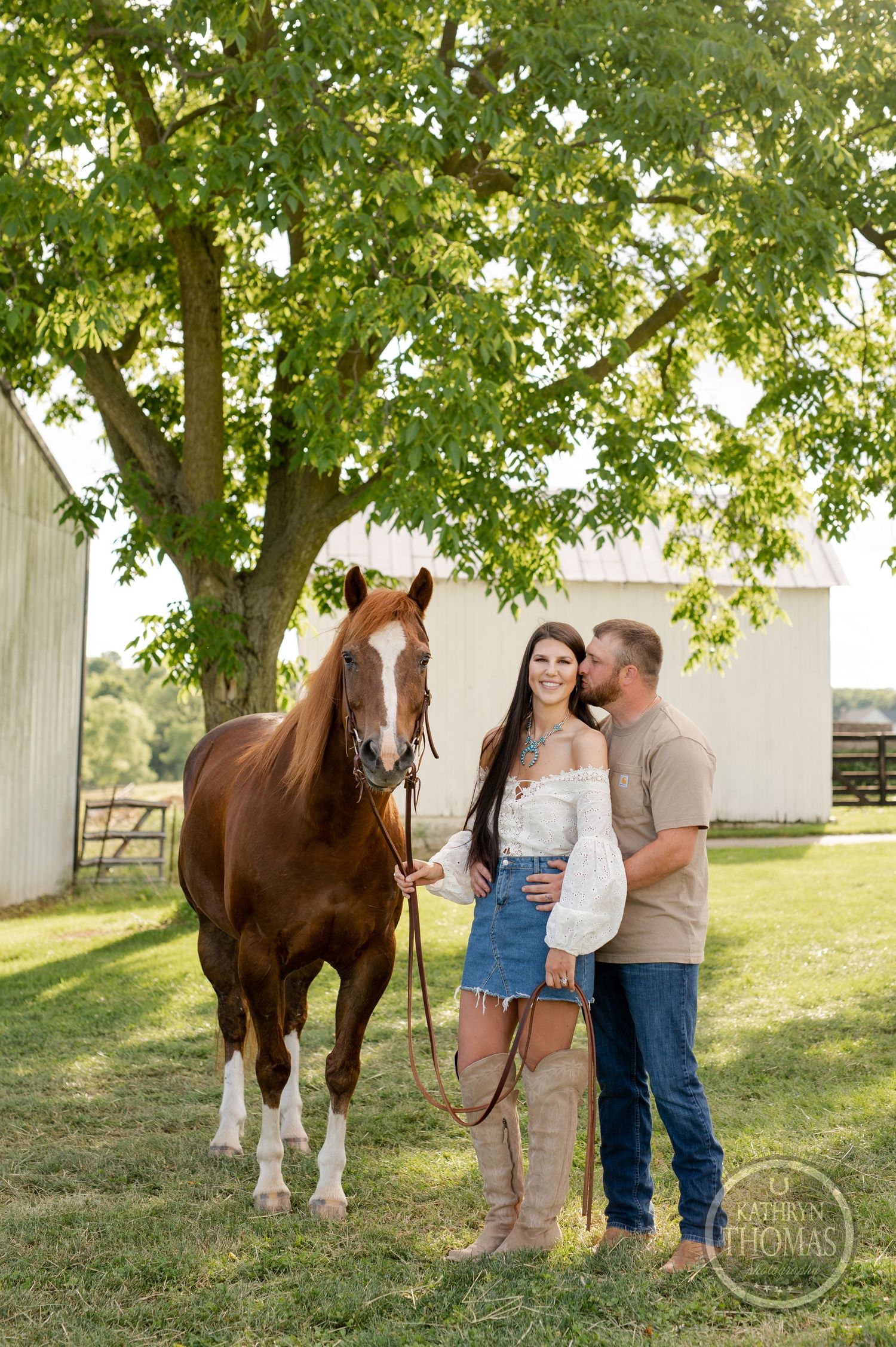 Couple poses with brown horse on grassy lawn near white barn structure.