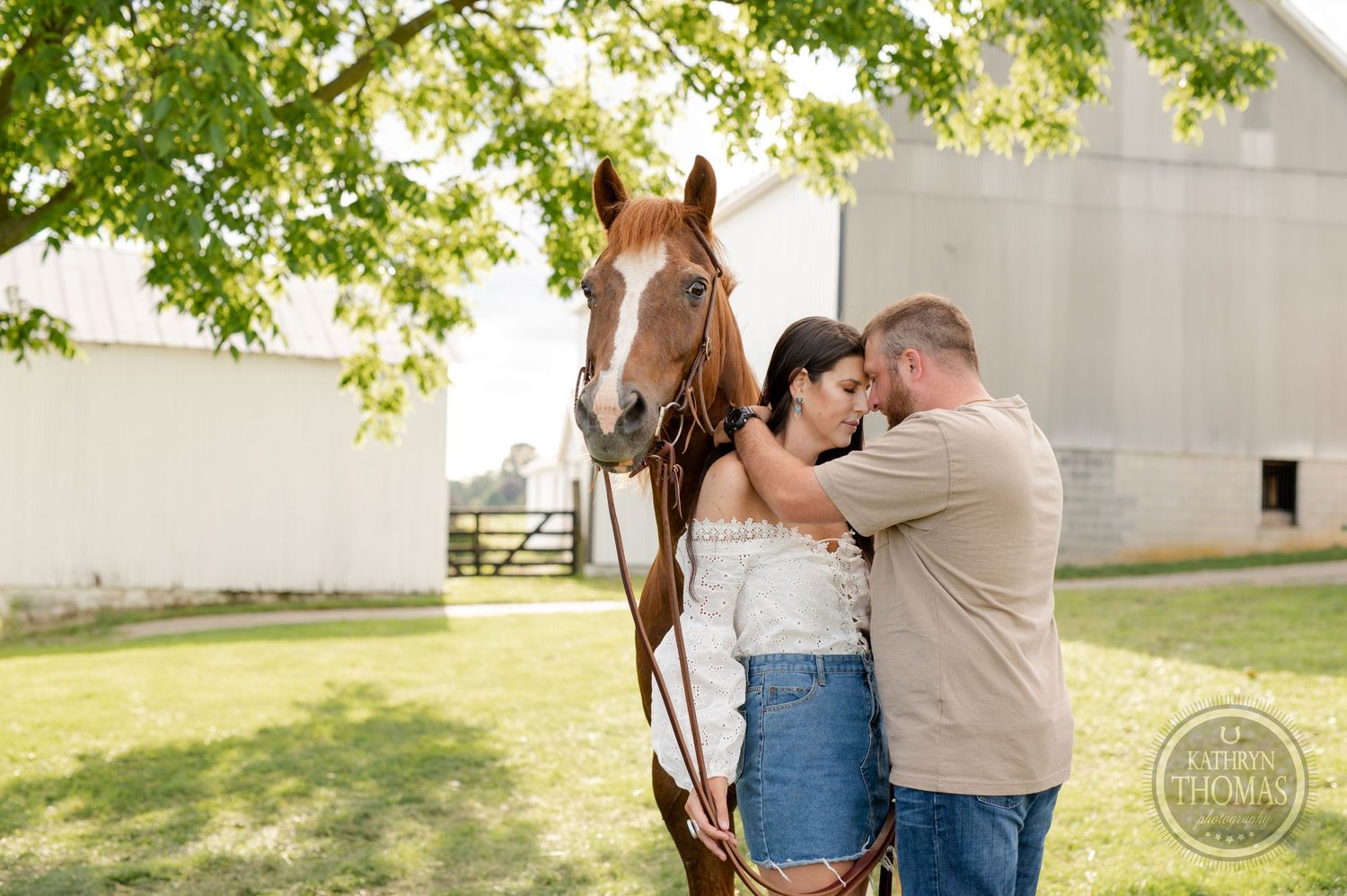 A romantic couple shares a tender moment in front of a horse on a sunny farm with white barn backdrop.
