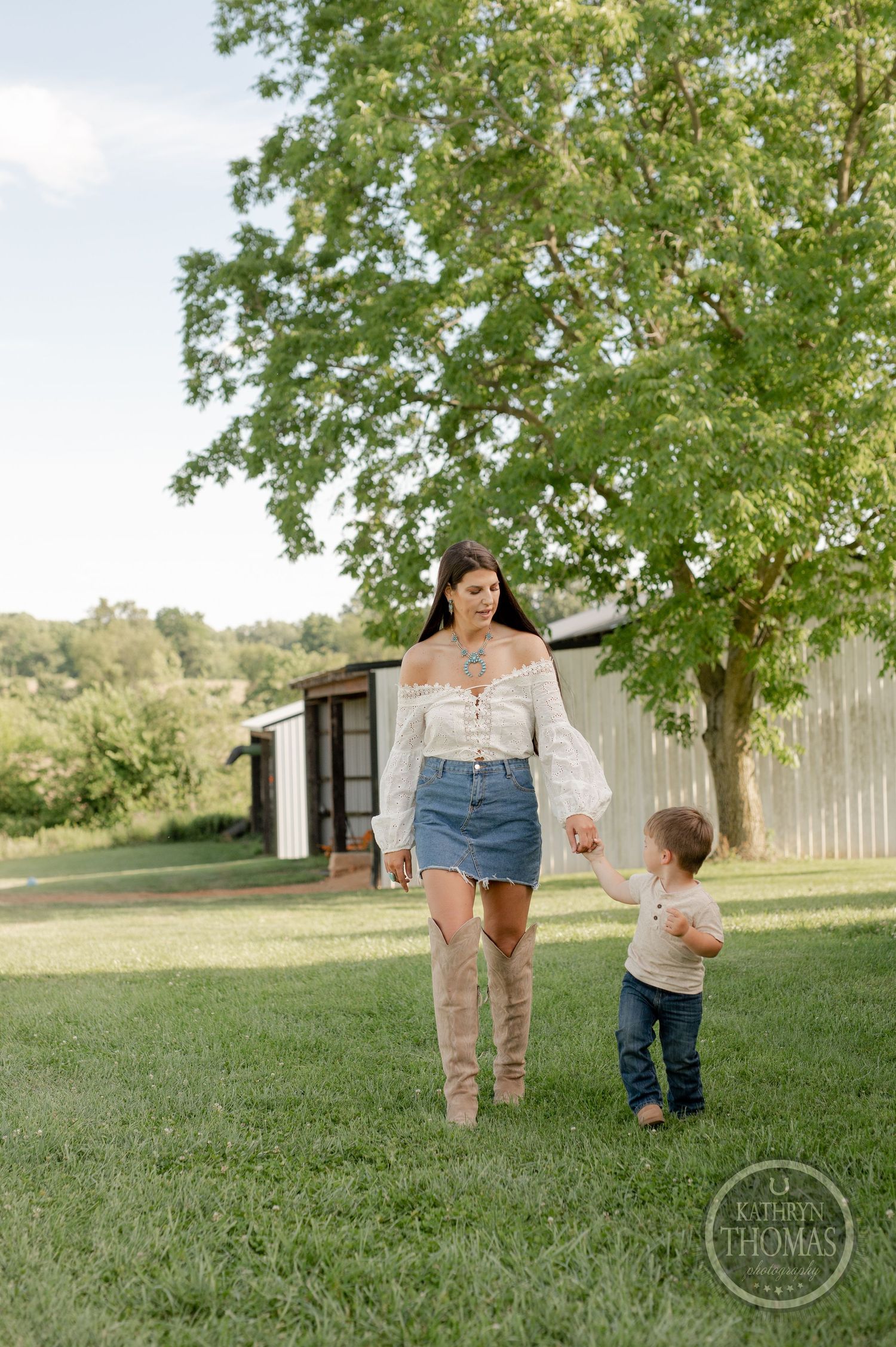 Woman in boots and child walking on grass near white building on sunny day.