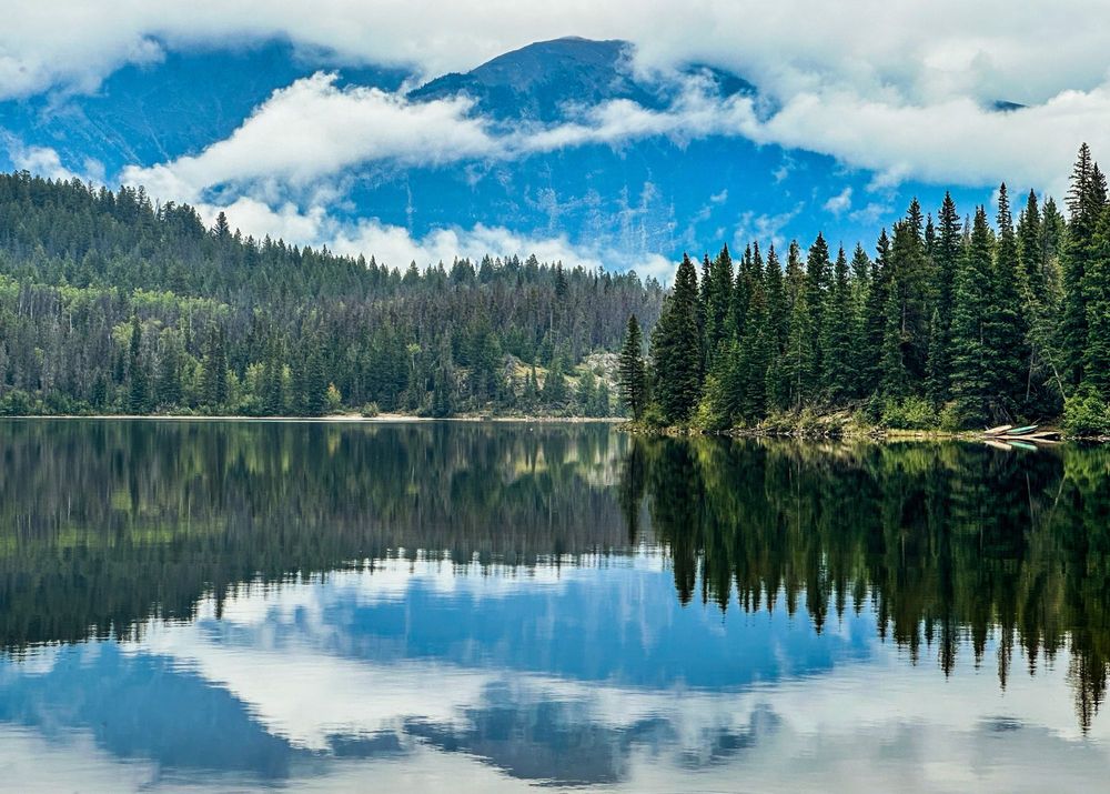 Mirror-like alpine lake reflects evergreen forest and puffy white clouds against vibrant blue mountain sky.
