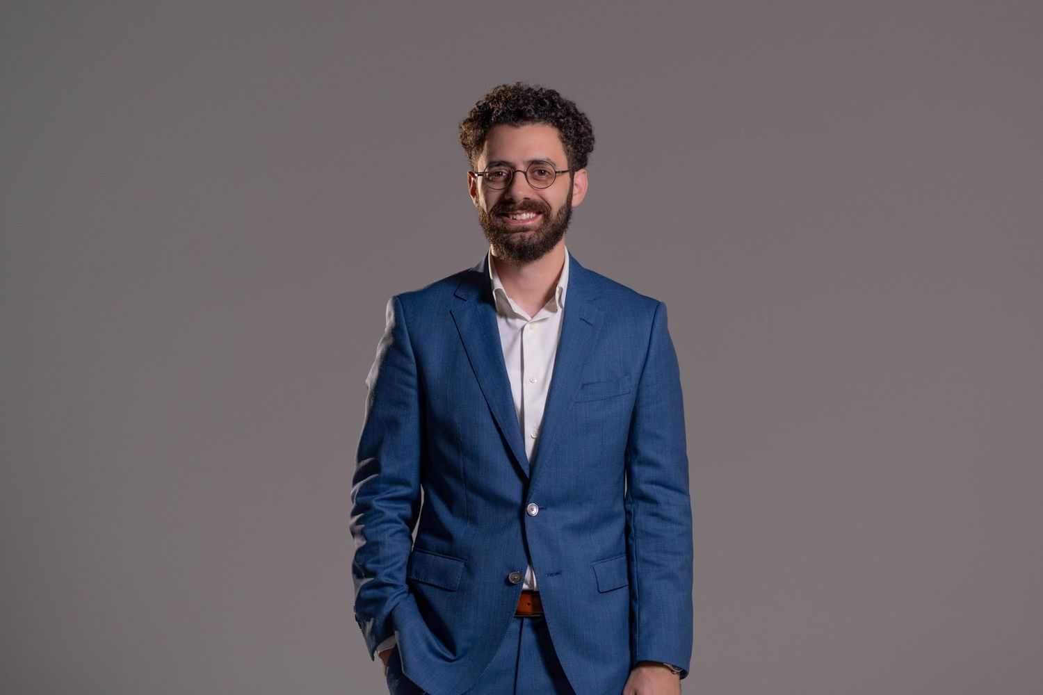 Professional portrait of person in blue suit jacket and glasses against grey background.