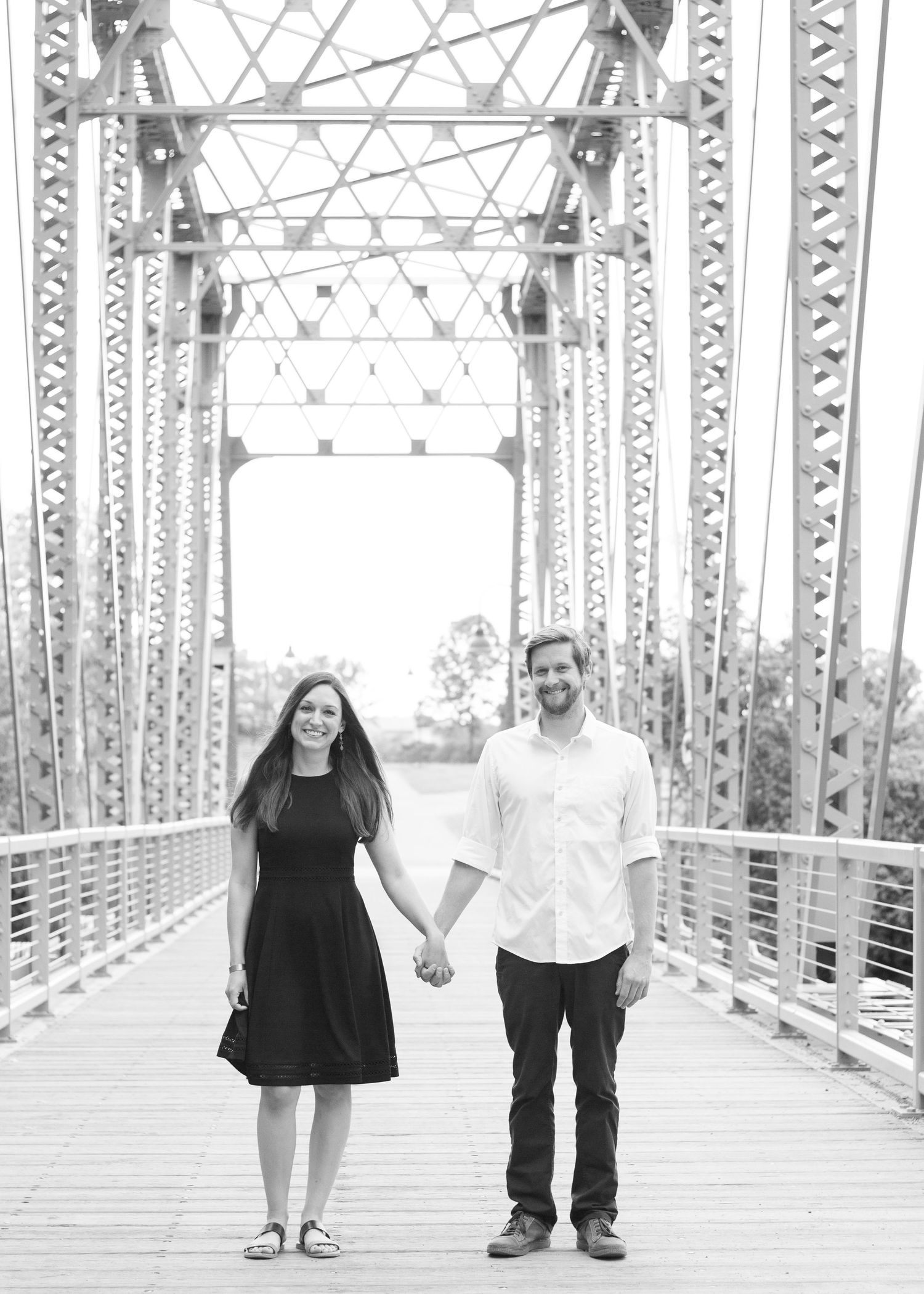 Couple holding hands while standing on vintage metal bridge in black and white photo.