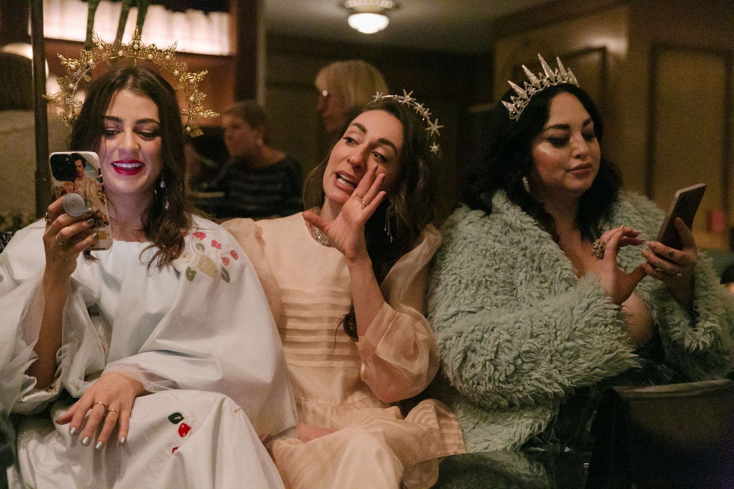 Three friends in wedding attire sit together on a couch while looking at their phones and laughing.