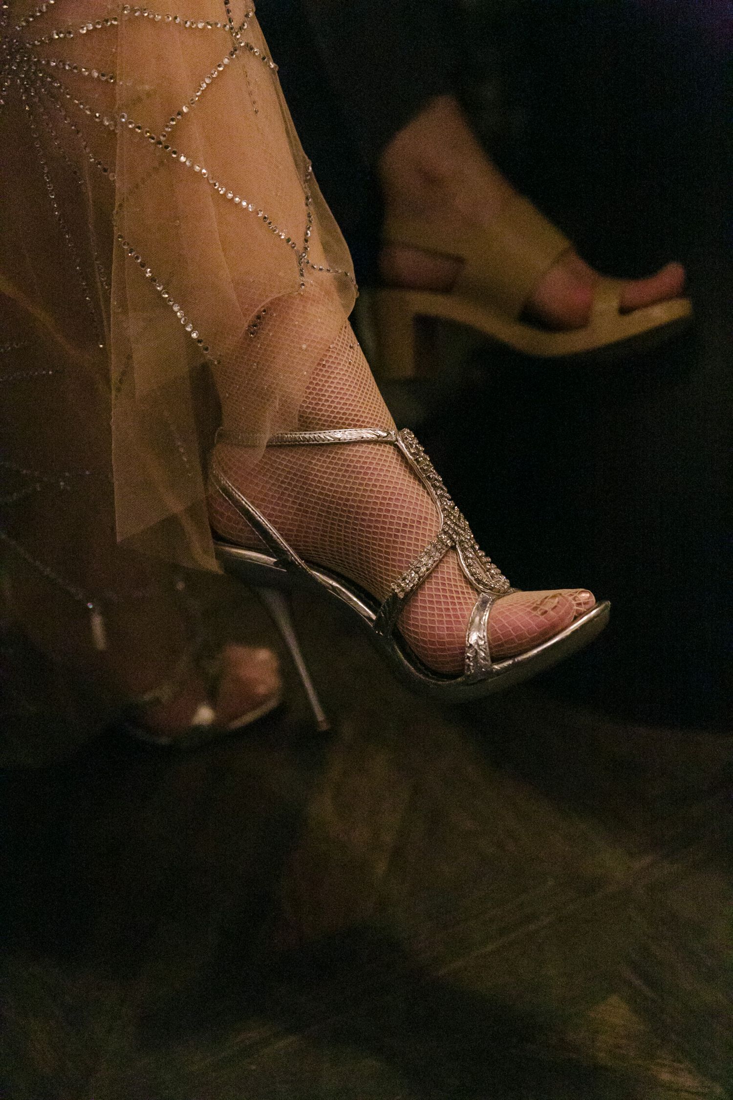 Elegant silver strappy high heel sandal with rhinestones shown through sheer fabric against dark background.