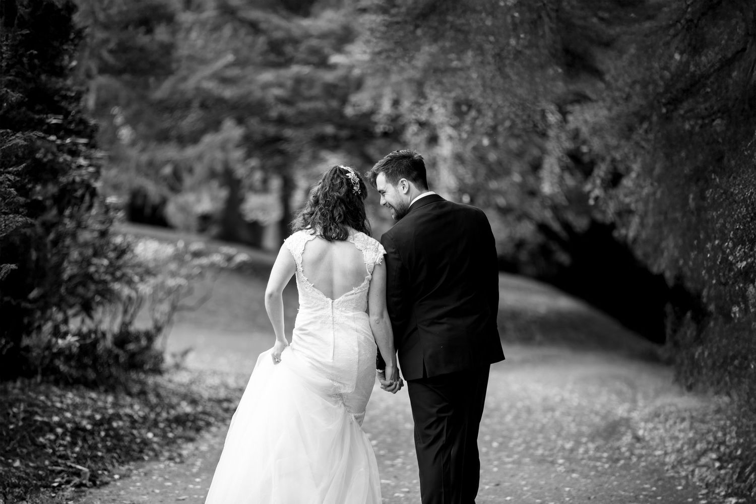 A romantic black and white photo shows a couple in wedding attire walking along a garden path surrounded by foliage.