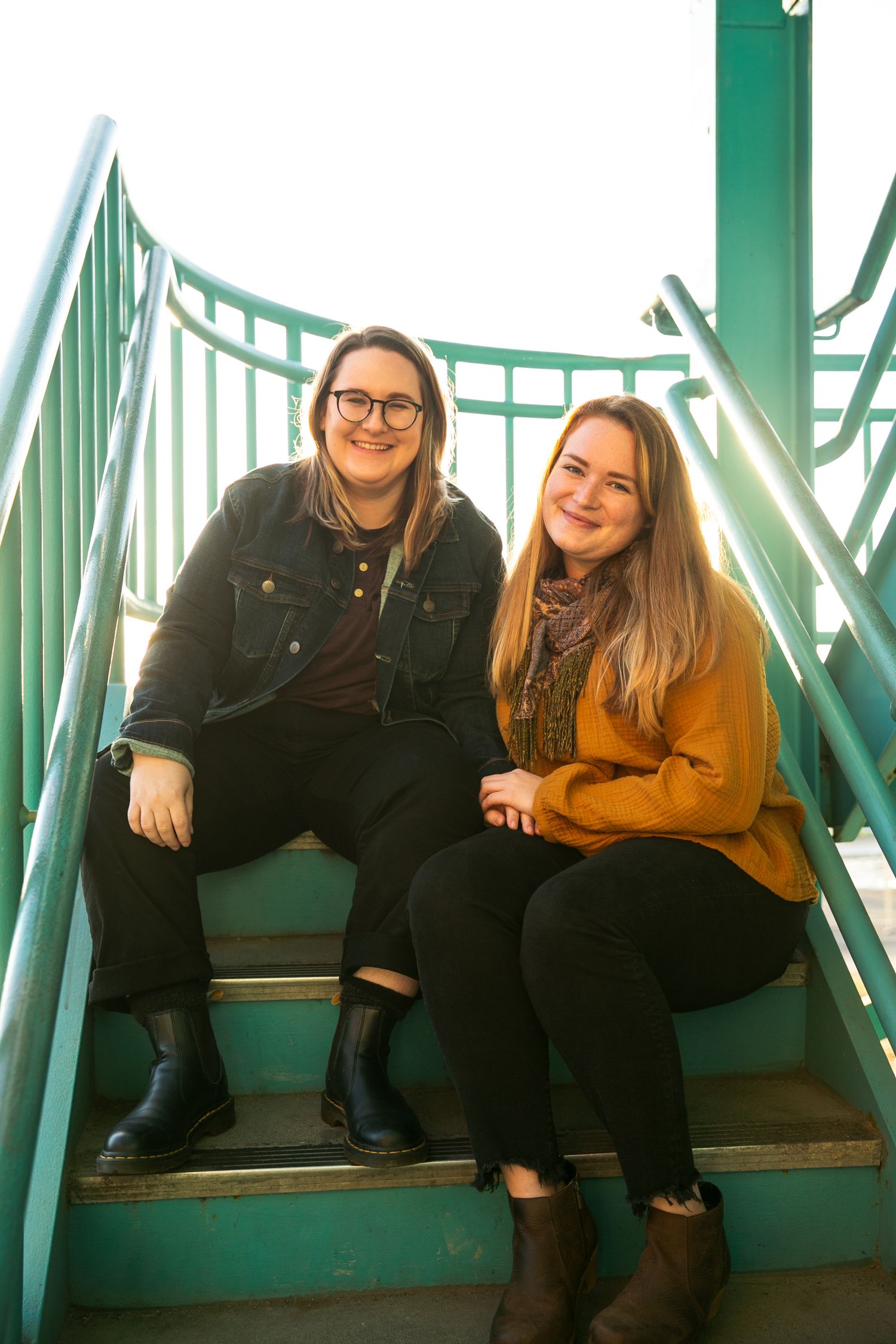 Two people sitting on bright green metal stairs wearing casual clothing and smiling at the camera.