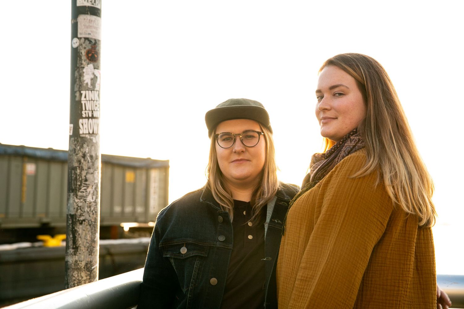 Two people in casual winter clothing stand together outdoors against a sunset-lit urban backdrop.