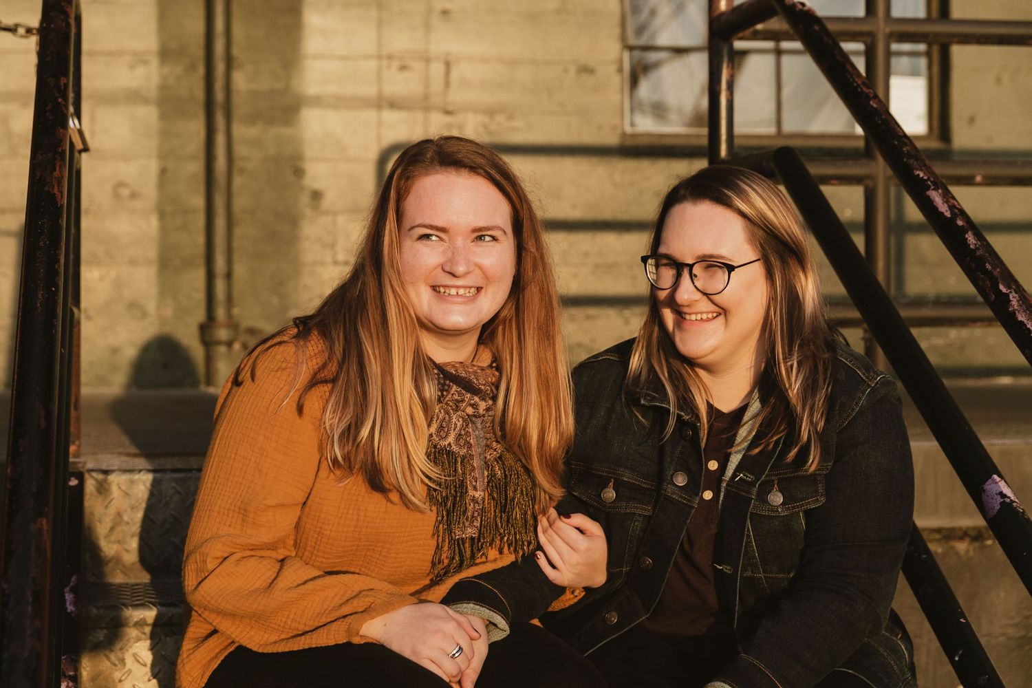 Two friends sitting together and smiling on stairs with a brick wall background during golden hour.