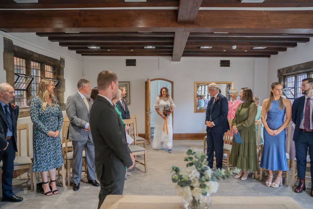 People gather for an intimate indoor wedding ceremony in a rustic room with wooden beams and natural lighting.