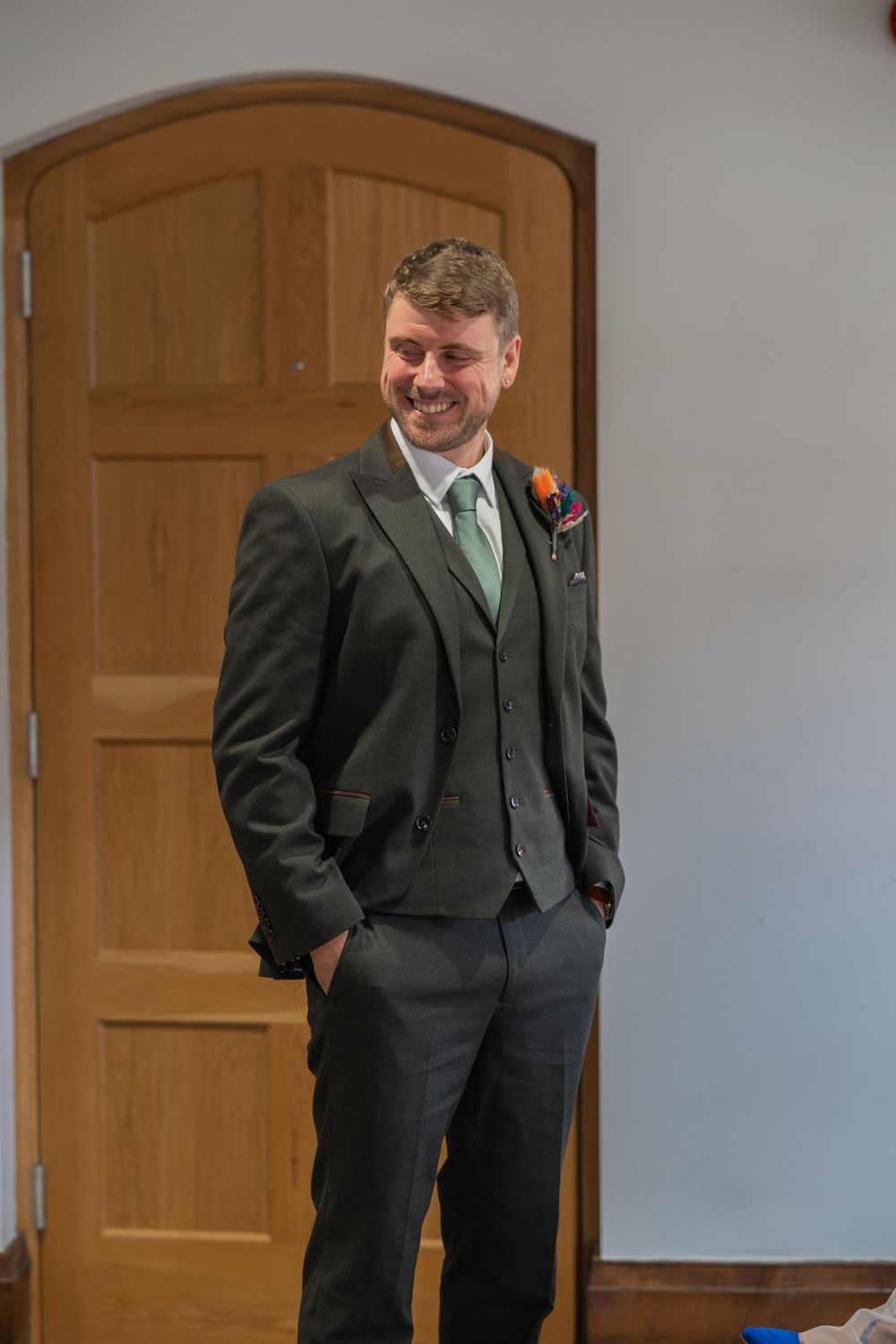 Someone wearing a dark three-piece suit with a green tie and boutonniere smiles in front of a wooden door.