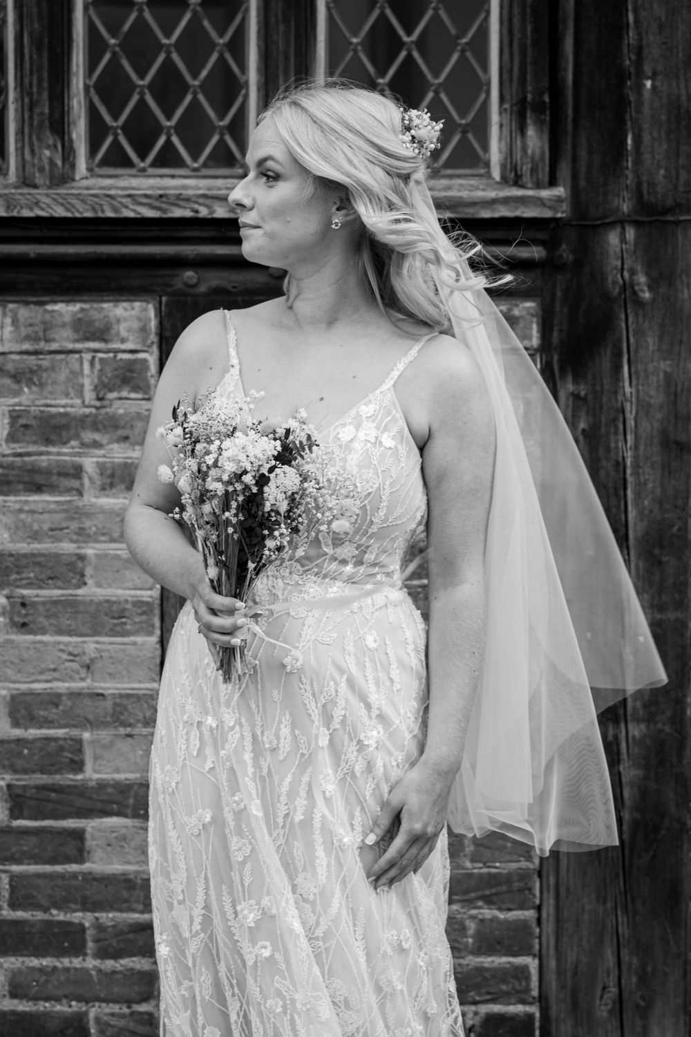 Black and white vintage-style bridal portrait with flowing lace dress and veil against a rustic brick wall background.