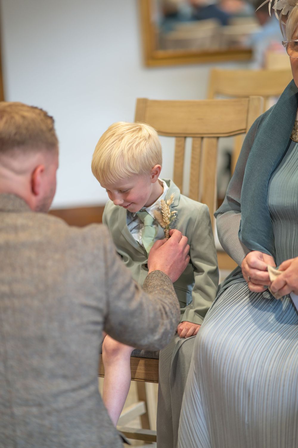 A young boy in a light suit has a boutonniere pinned to his lapel during a formal occasion or wedding ceremony.