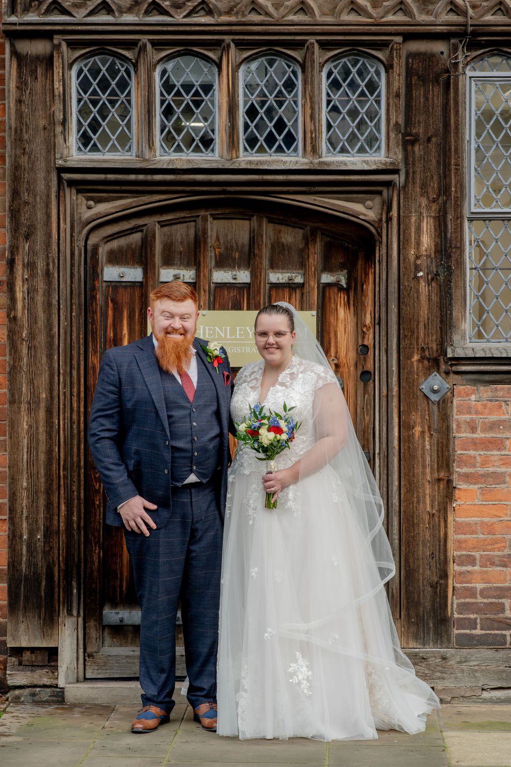 Wedding couple poses in front of ornate wooden Tudor doorway with diamond-paned windows and brick walls.