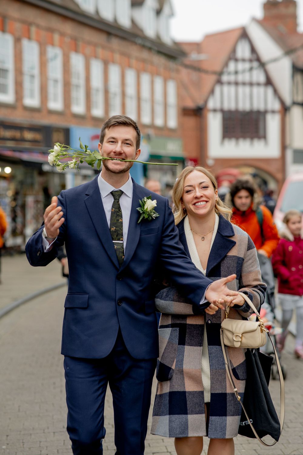 A couple walking down a charming street with Tudor-style buildings wearing matching navy blue formal attire.