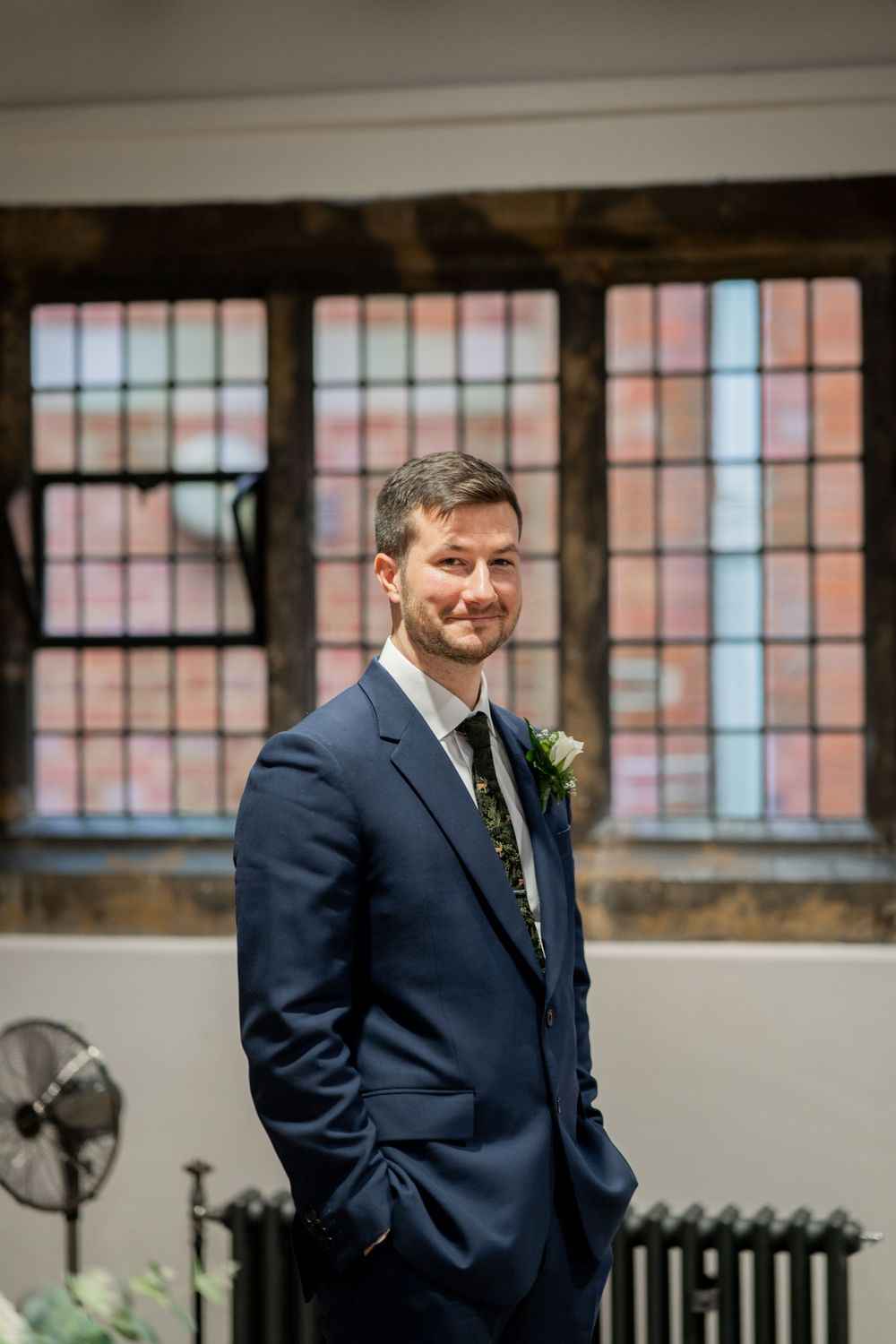 A groom in a navy blue suit stands smiling indoors near windows with a boutonniere pinned to his lapel.