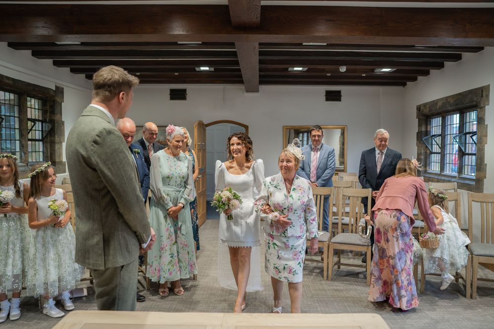 A group of elegantly dressed wedding guests gather in a rustic room with exposed wooden beams and large windows.