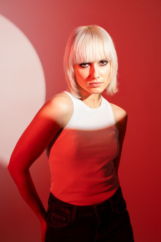Person with platinum blonde hair poses against red background with dramatic lighting.
