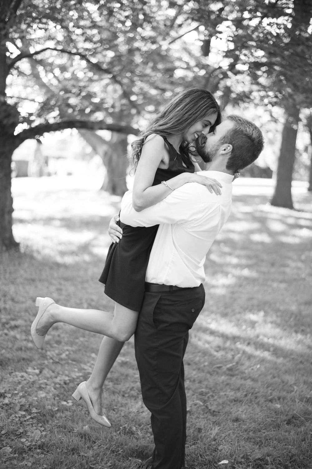A black and white photo of a couple sharing a playful embrace in a tree-lined park setting.