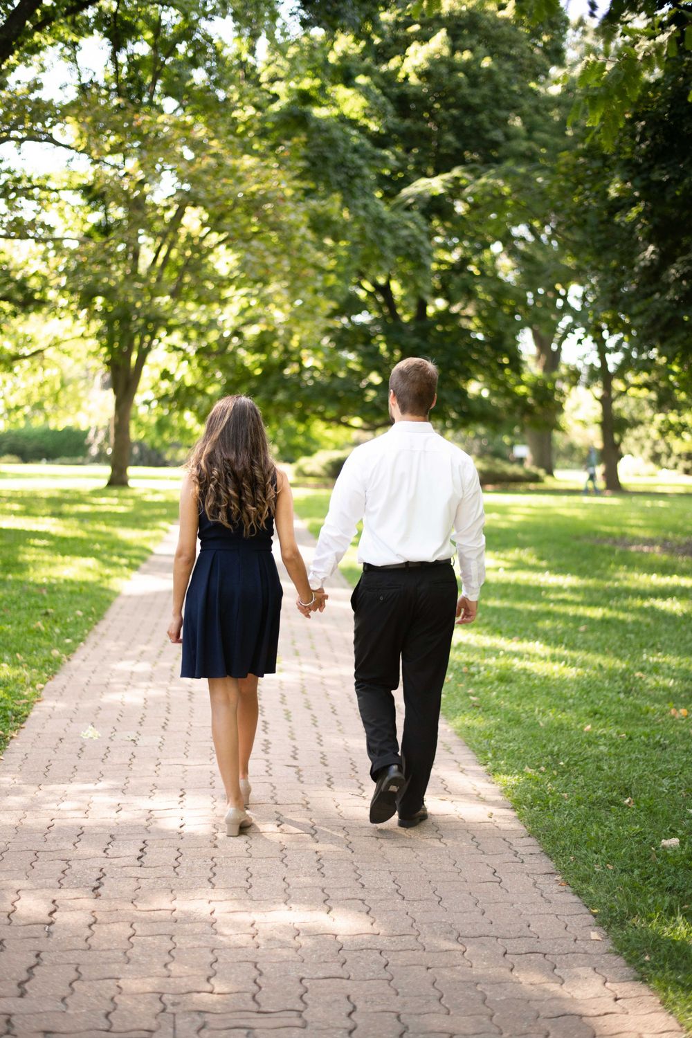 A couple holds hands while walking down a brick path surrounded by lush green trees in a summer park.