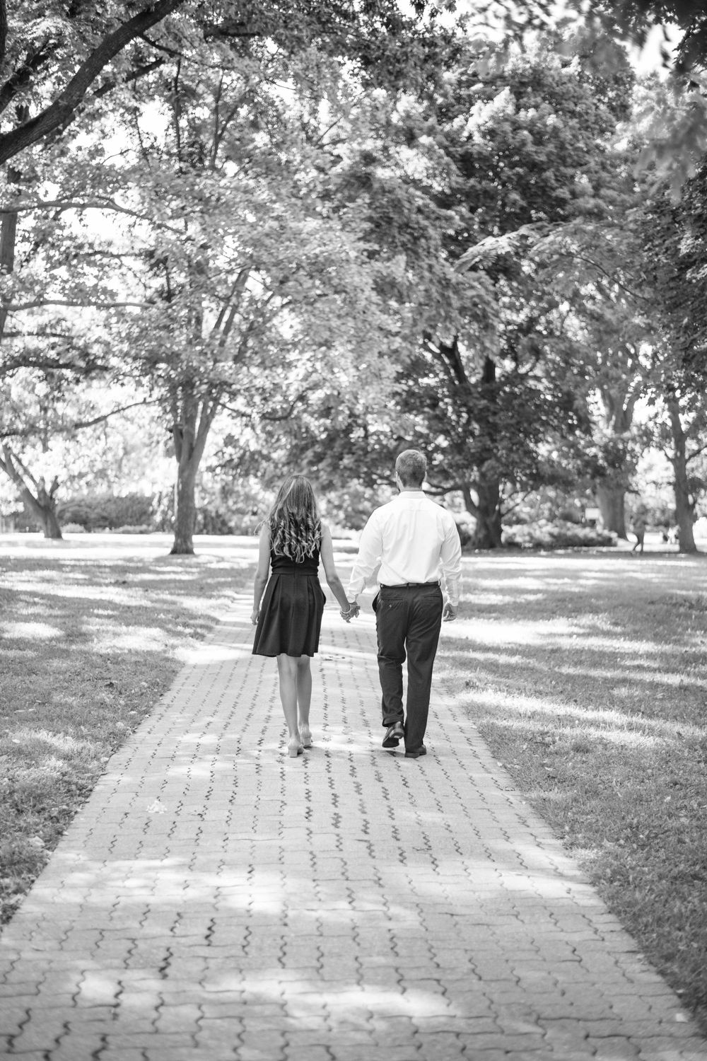A couple holding hands while walking along a brick pathway surrounded by trees in a peaceful park setting.