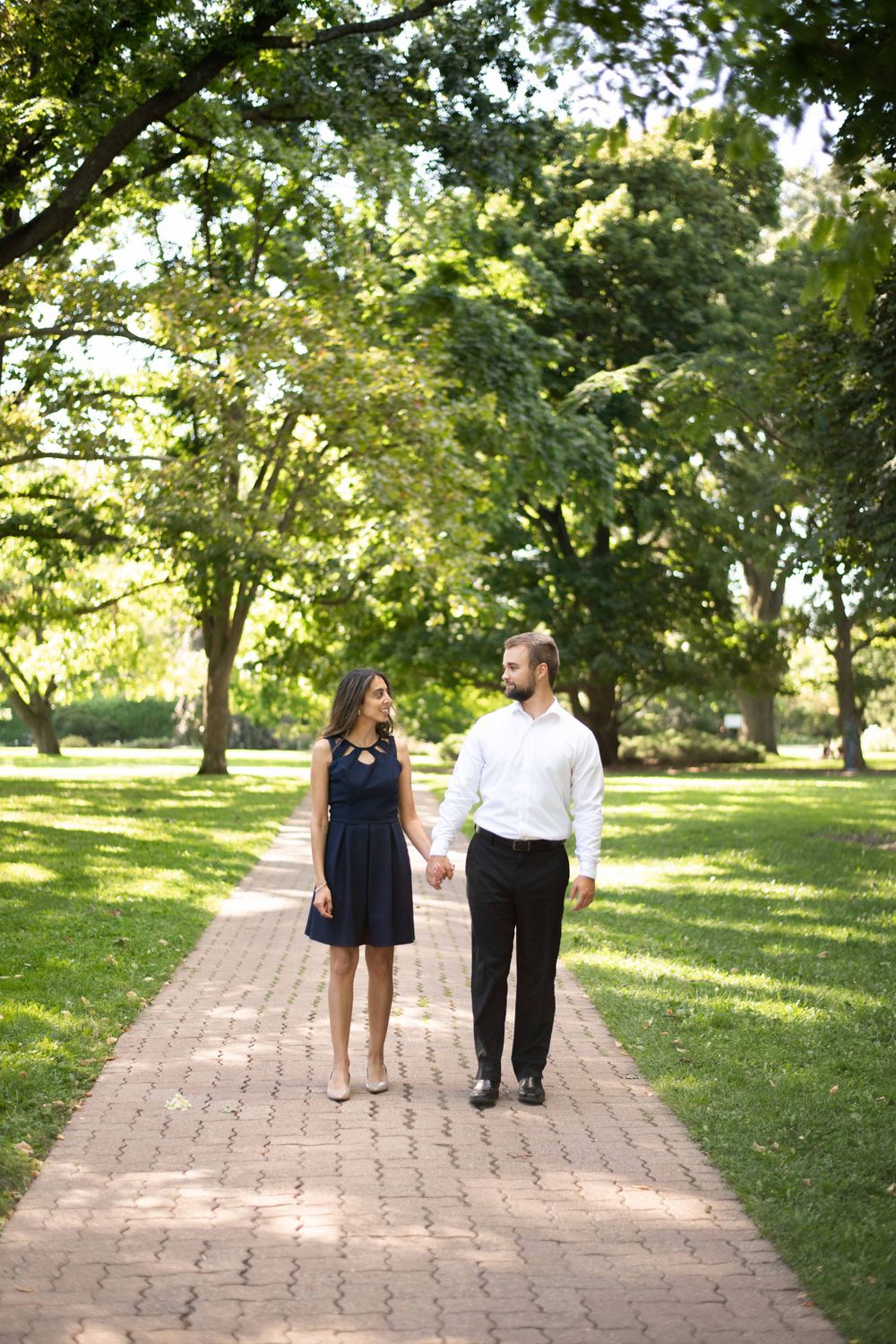 A couple walks hand in hand along a brick path through a sunny green park on a summer day.
