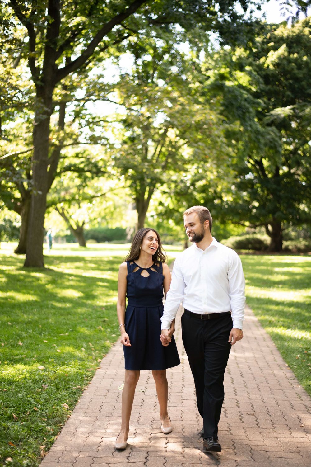 A couple walking together on a brick path through a sunny park with green trees and grass.