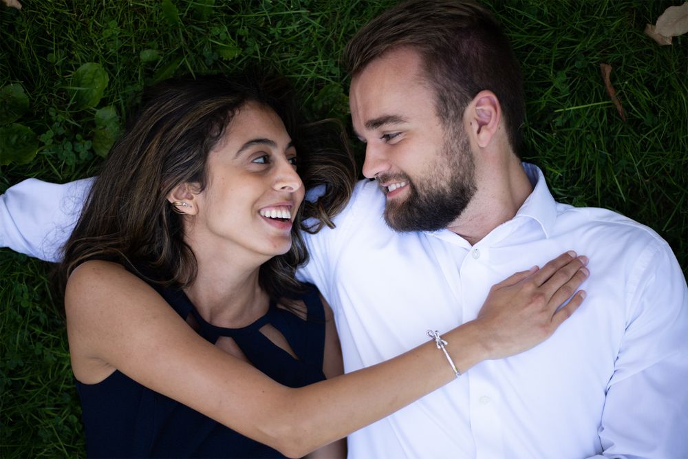 A couple lies together on green grass, sharing a joyful moment while wearing navy blue and white casual attire.