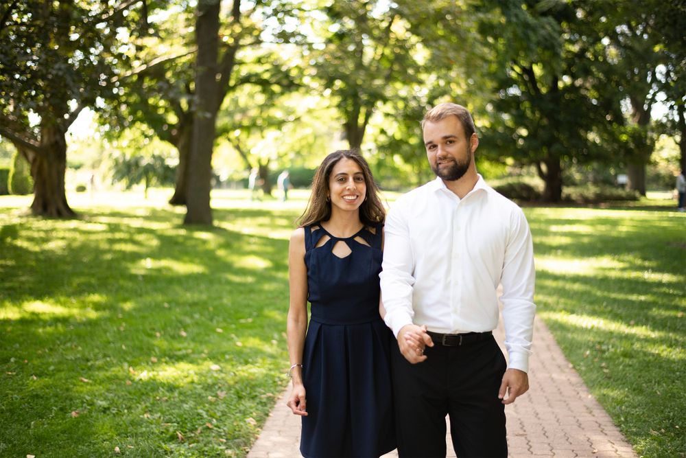 A couple holding hands walks along a sunlit garden path surrounded by lush green trees on a summer day.