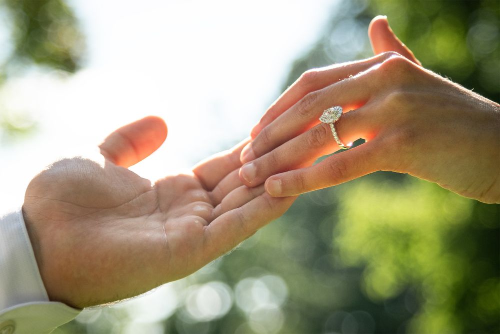 Two hands tenderly touch as a diamond engagement ring sparkles in soft natural light with bokeh background.