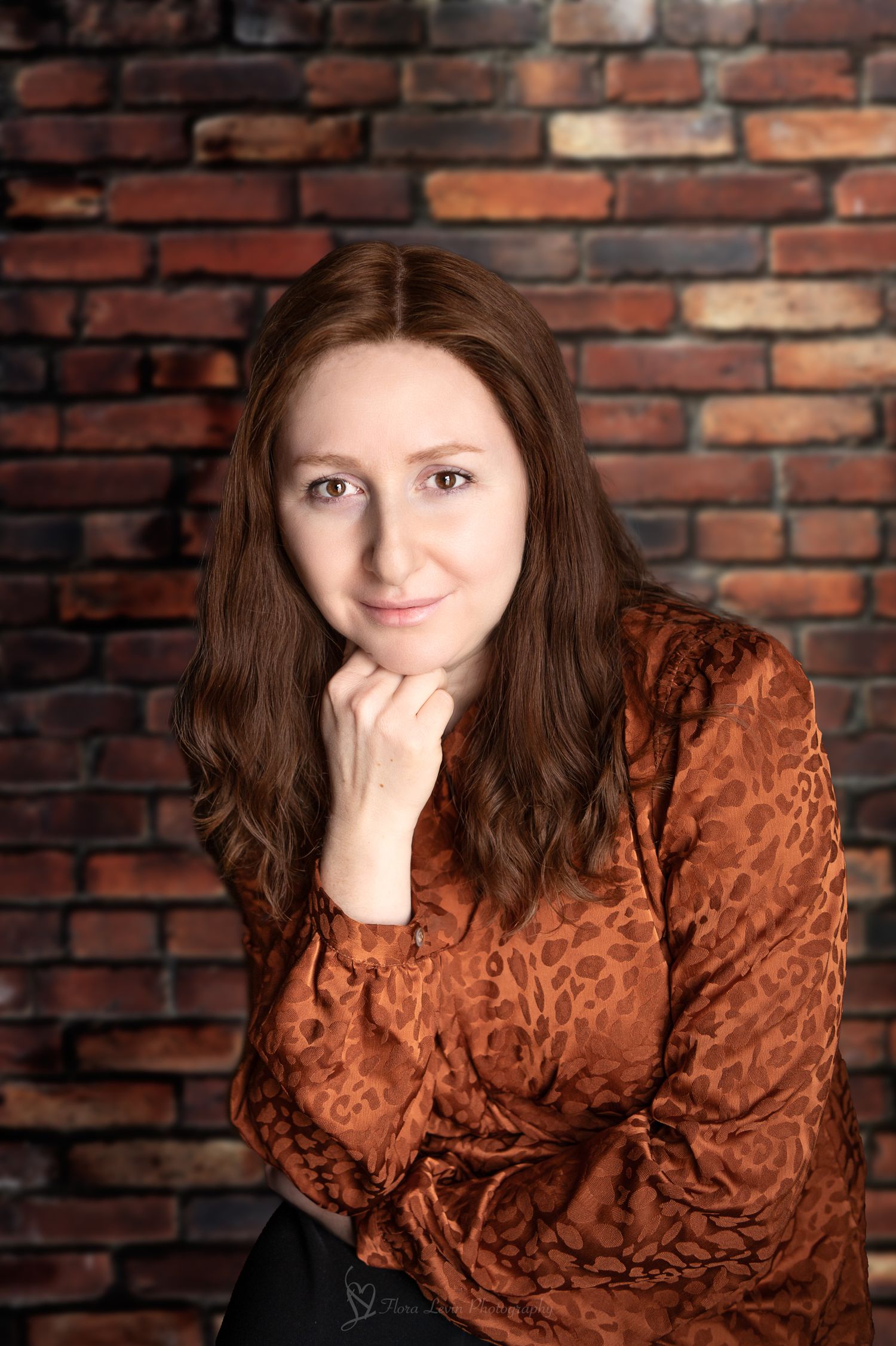 A woman in a copper-colored satin top poses against a rustic brick wall with a thoughtful expression. Photo  by Flora Levin Photography.