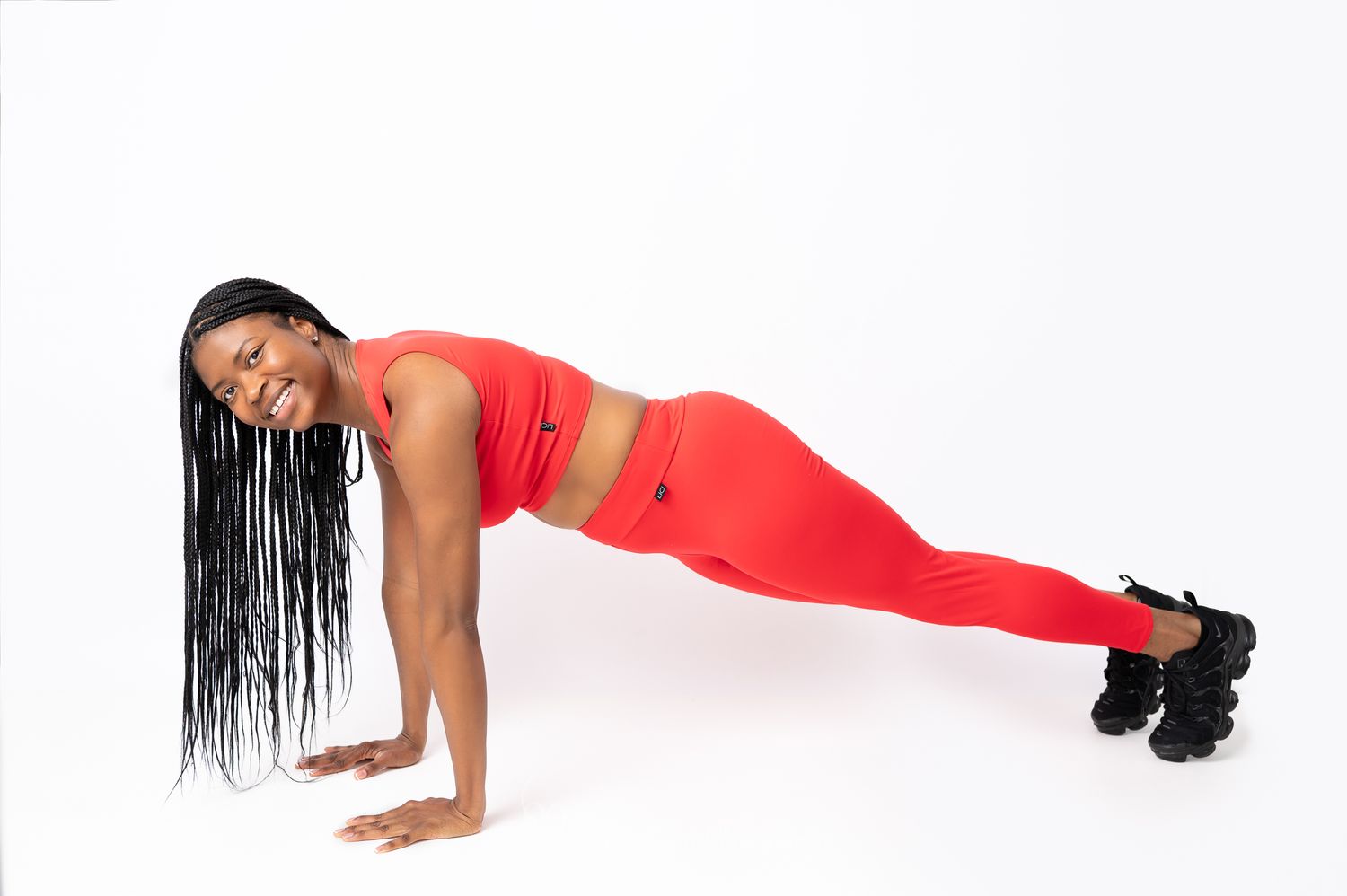 A personal trainer in a red athletic outfit demonstrates a plank exercise position on a white background.