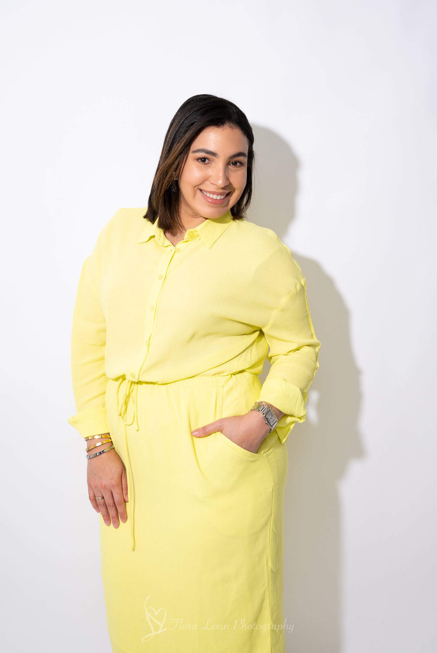 Woman wearing a bright yellow long-sleeved dress with a tie waist poses against a white background. In a sunny lit studio lighting.