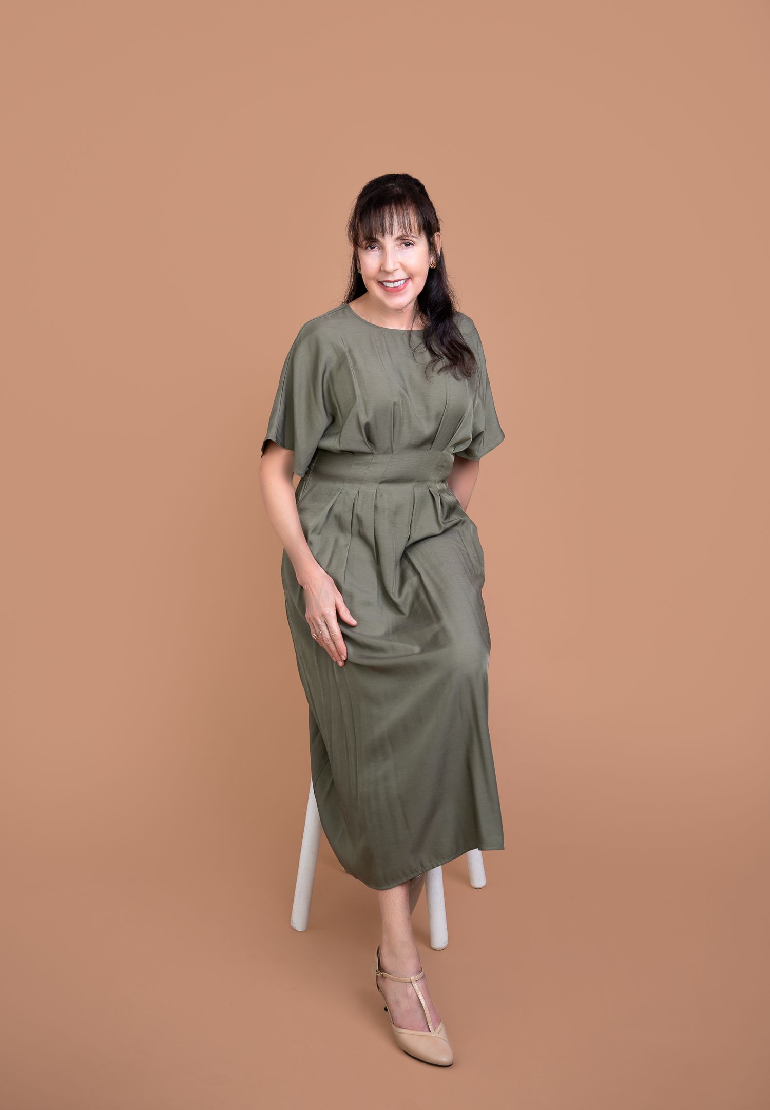 Woman poses for personal branding photoshoot in an olive green ruffle dress with beige shoes against a neutral background.
