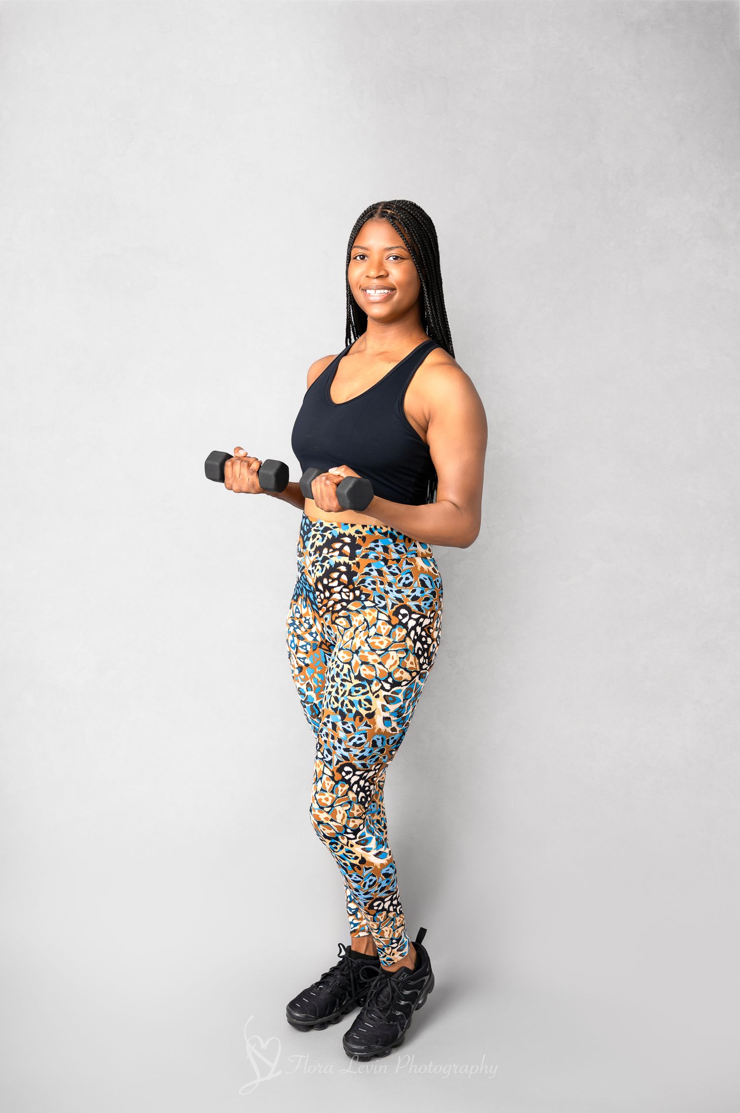 A personal trainer wearing a black tank top and leopard print leggings poses holding weights. In a plain backdrop.