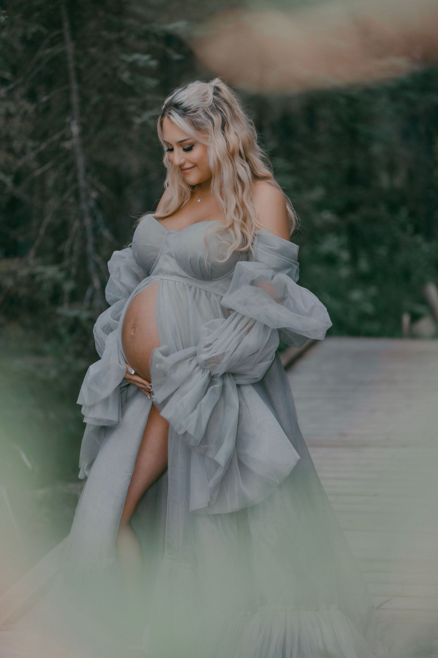 Ethereal maternity portrait in flowing grey dress against moody forest backdrop with dreamy lighting effects.
