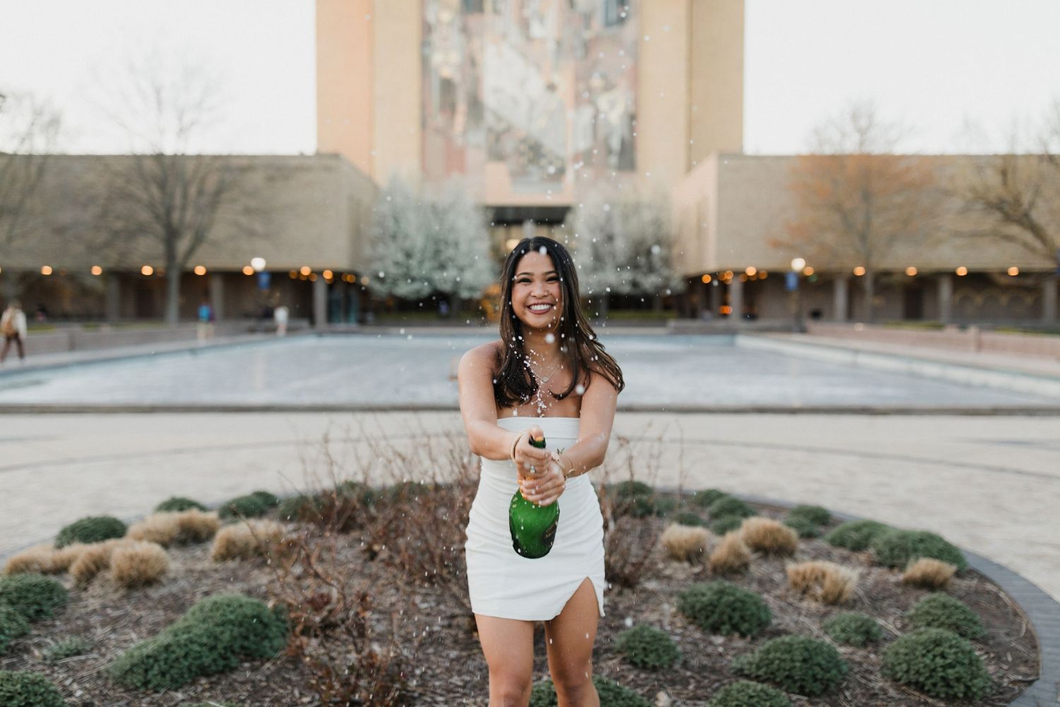 Graduate in white dress holding a celebratory drink outdoors with architectural backdrop.