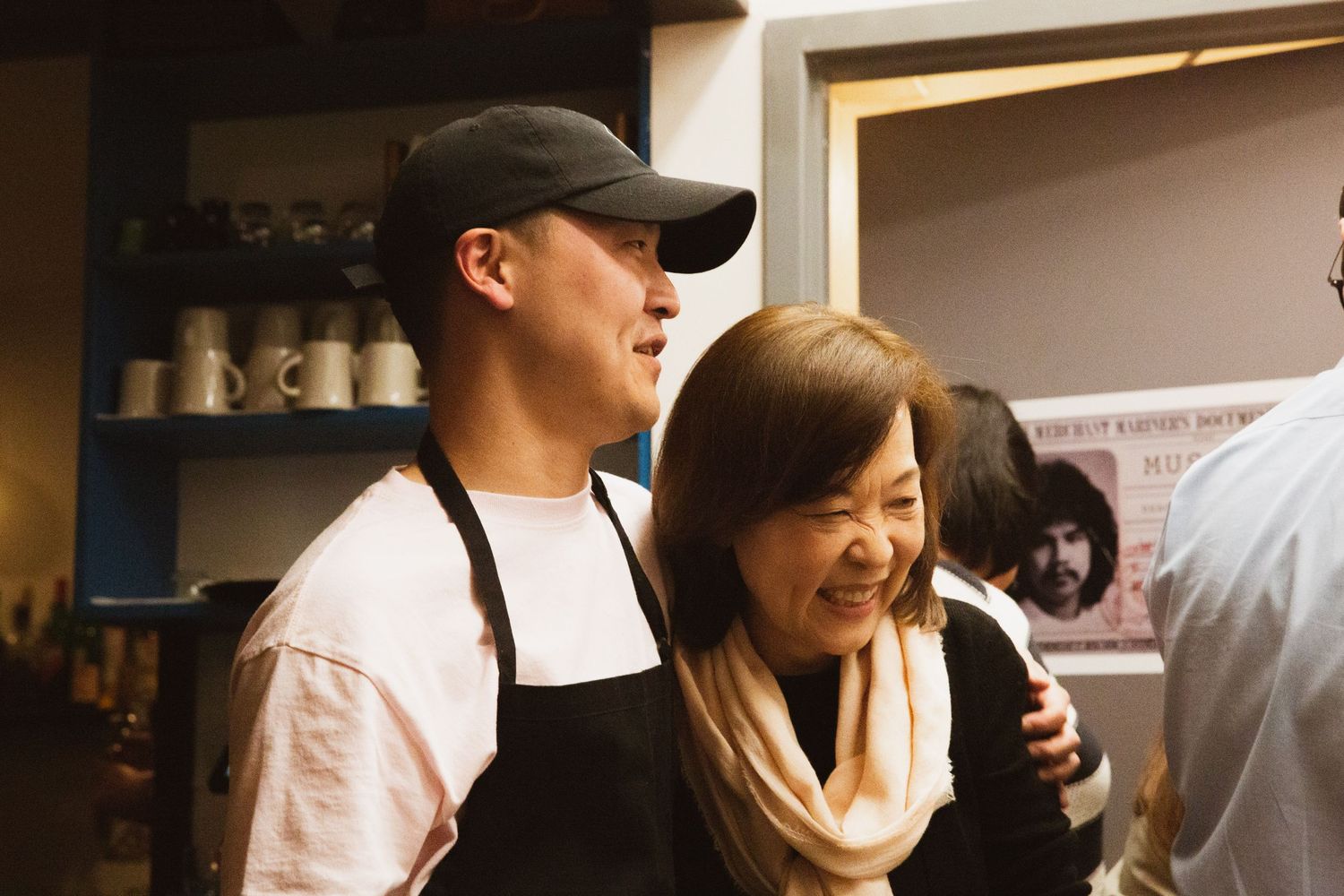 Two people working in a restaurant kitchen smile together in a candid moment, with storage shelves visible in the background.