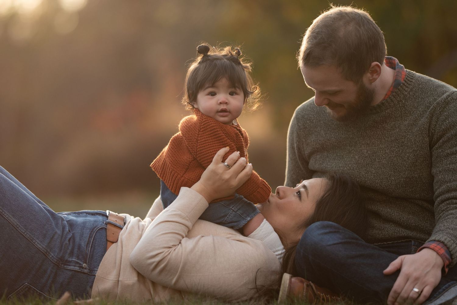 Young couple lying in autumn grass smiling at their baby girl in a warm fall outdoor setting. Photo by Xenophon Photography in Maine.