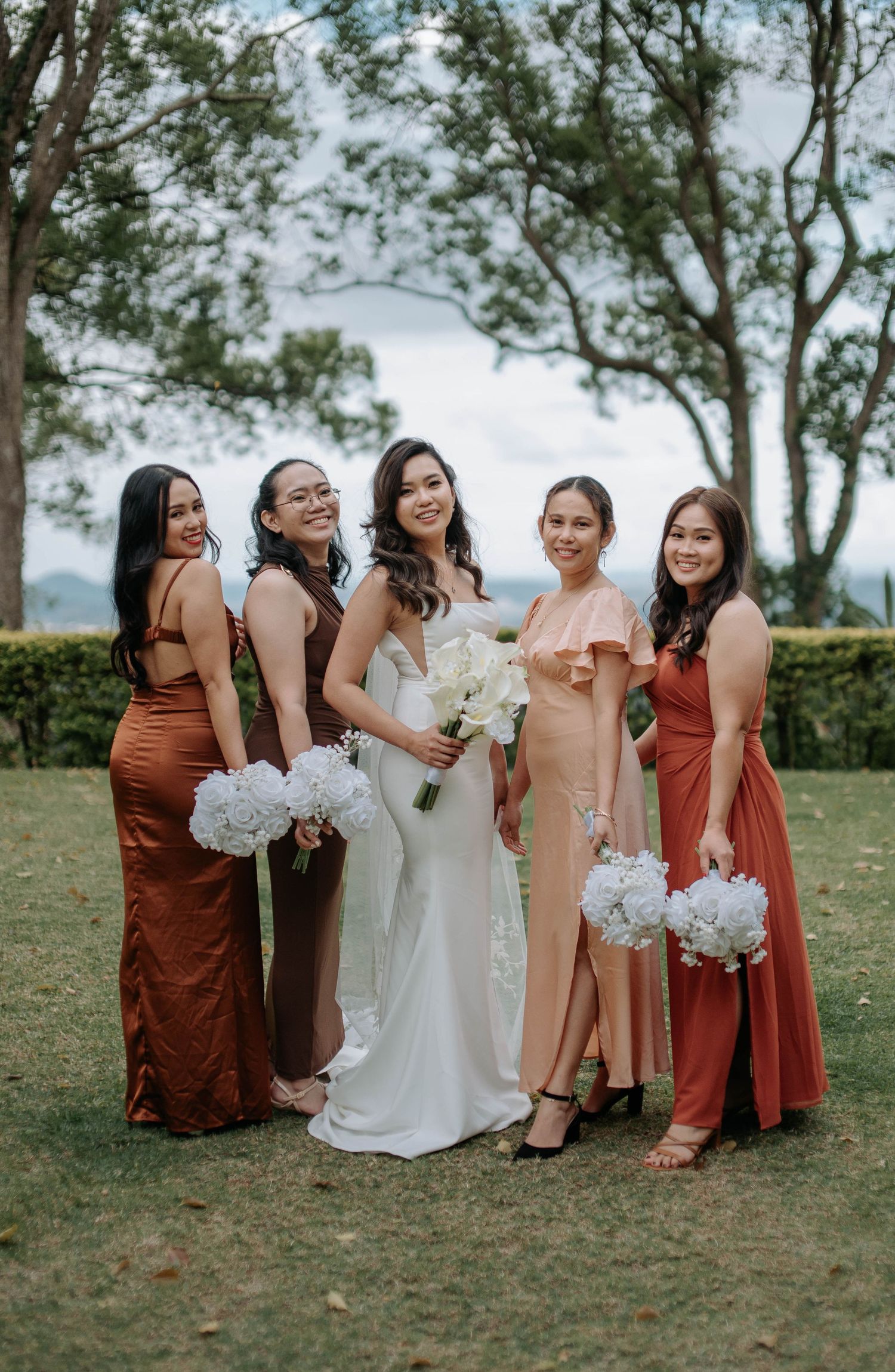 Wedding party in terracotta and rust-colored dresses pose together on grassy lawn during outdoor celebration.