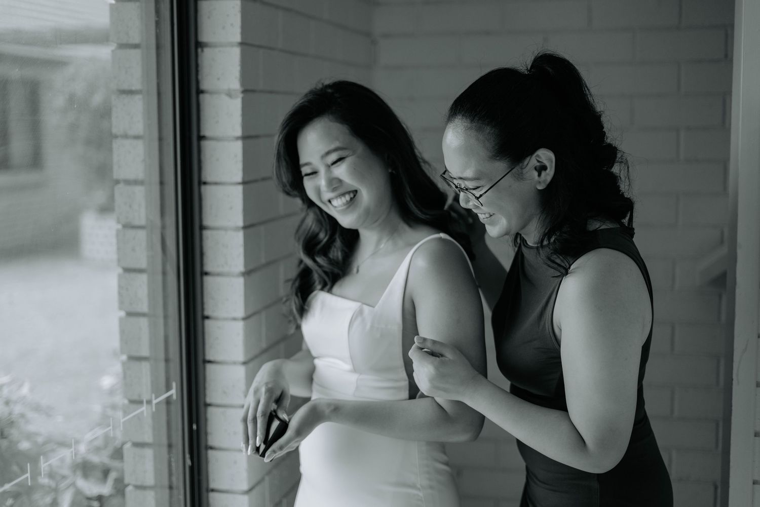 Two friends share a joyful moment together in black and white, laughing by a window.