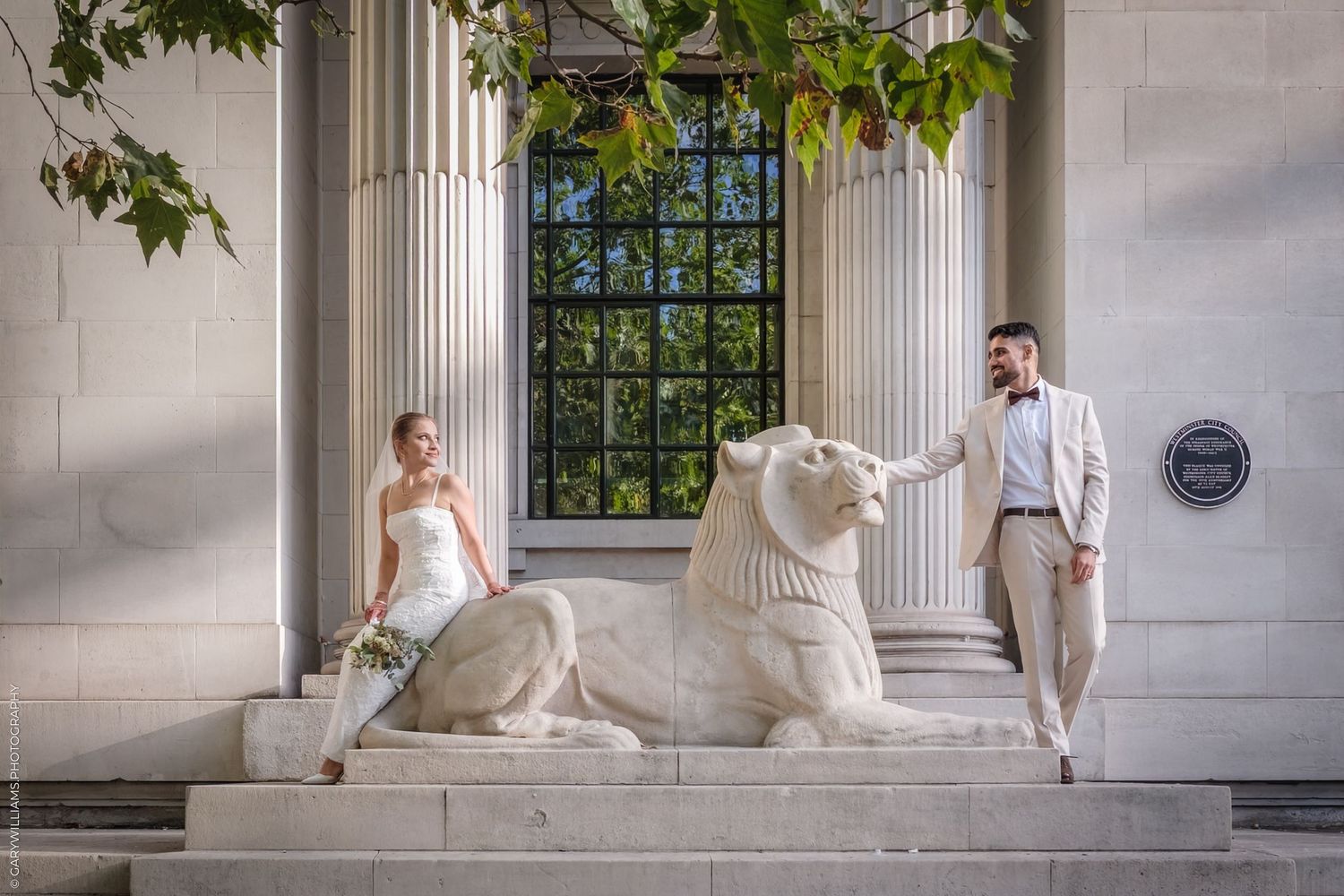 A couple shares a romantic kiss wearing formal attire during their wedding photos against a lush garden backdrop.