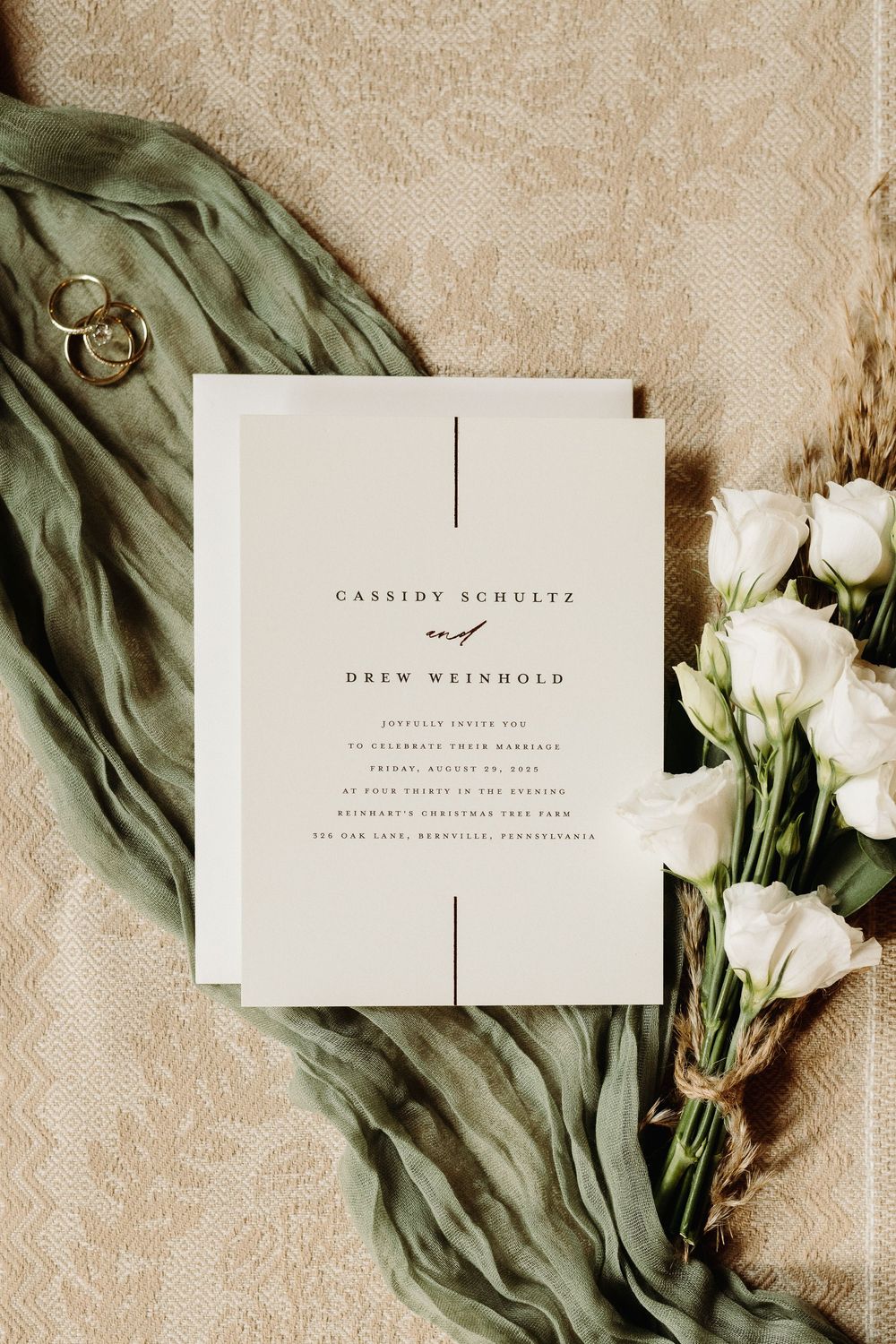 Elegant wedding invitations with green silk ribbon and white roses laid on beige textured background.