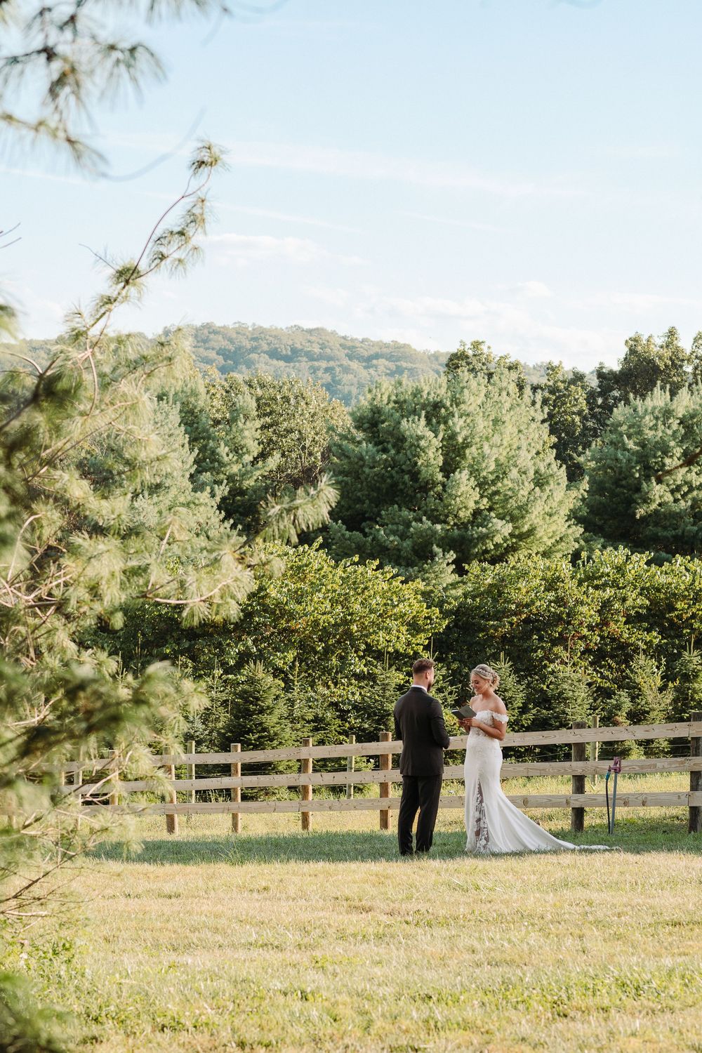 A couple stands together in an outdoor field with white fencing and trees in the background on a sunny day.
