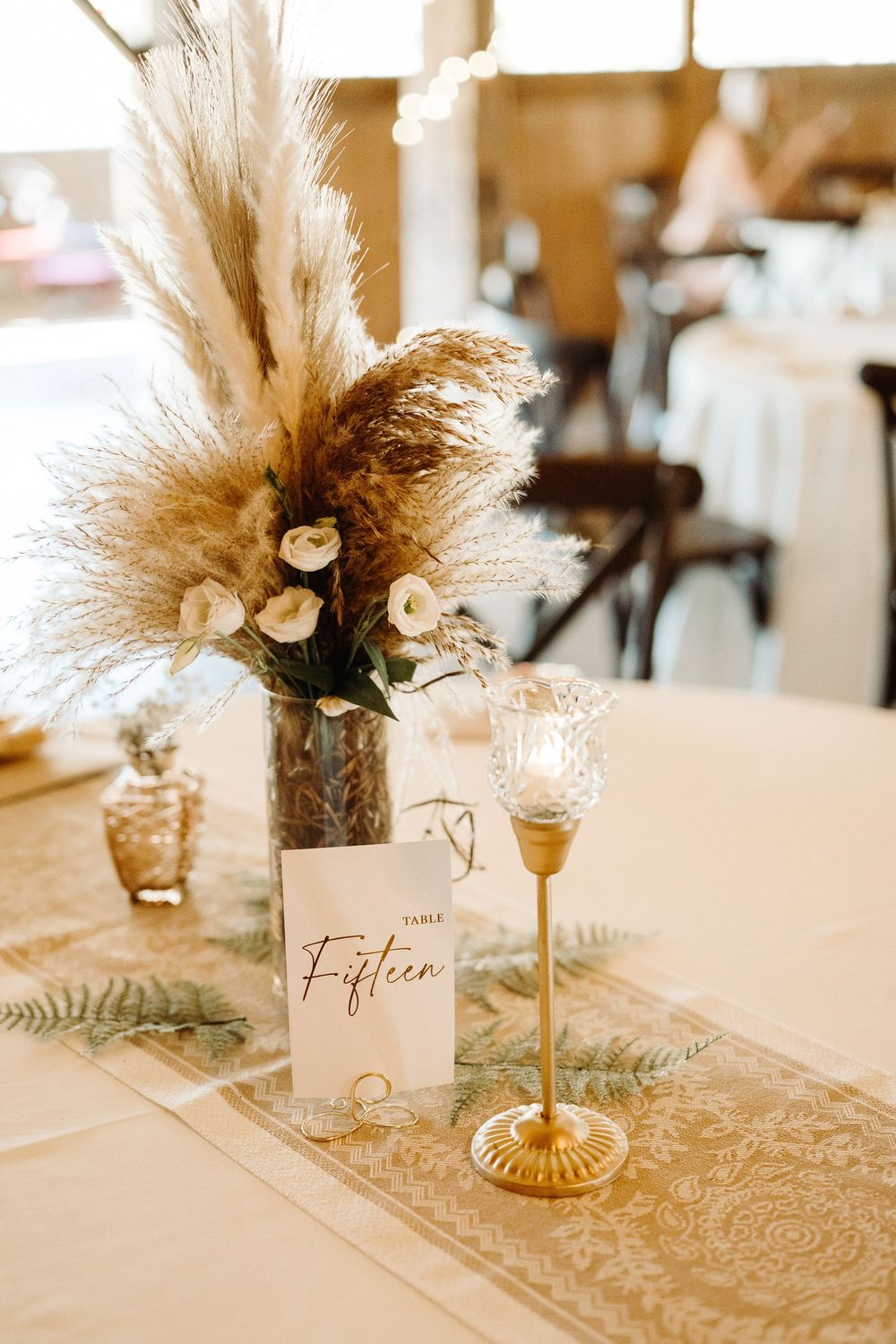 Elegant wedding table decor featuring pampas grass centerpieces and gold table numbers on white linens.