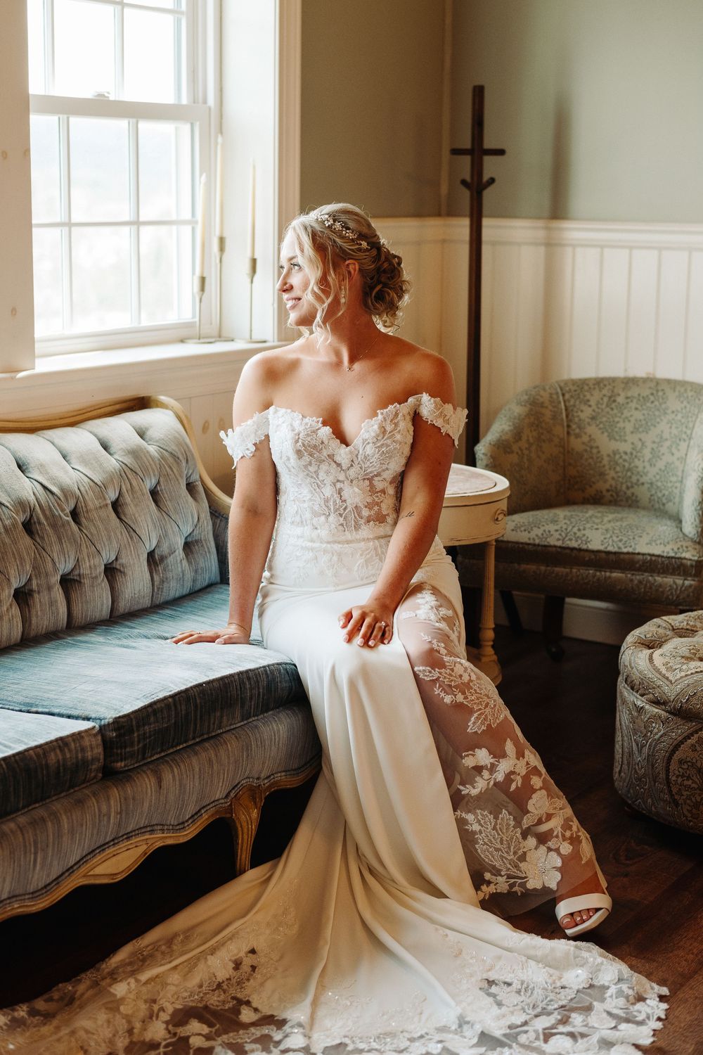 A beautiful white lace wedding dress with intricate details displayed on a gray vintage couch in natural window light.