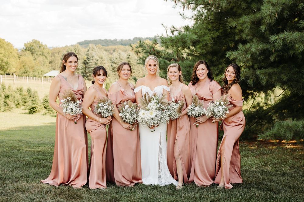 Bridesmaids in dusty rose dresses pose with bride in white gown outdoors for formal wedding photos.