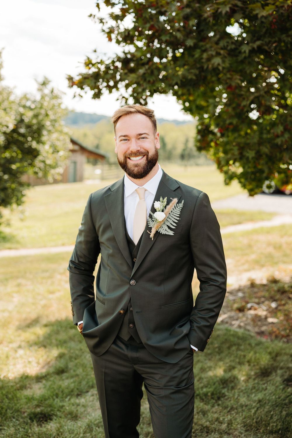 A groom in a classic black suit with a boutonniere poses outdoors against a natural backdrop.