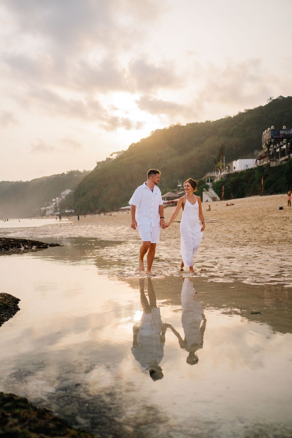 A couple in white clothing creates reflections while walking on wet beach sand at sunset.